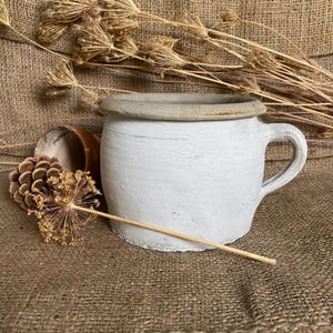 May include: A white ceramic mug with a handle and a light grey rim. The mug is set against a burlap background with dried plants and a small brown pot. The mug has a textured, painted surface.