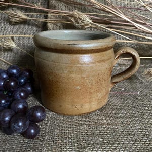 May include: A rustic, tan ceramic mug with a rounded body and a curved handle. The mug has a wide rim and a slightly textured surface. A bunch of dark purple grapes sits to the left of the mug. Dried plants are in the background.