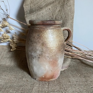 May include: An antique stoneware jar with a handle and lid. The jar has a mottled, earthy colour palette of brown, beige, and hints of pink. The surface has a textured appearance, suggesting age and use. The jar is likely used for storage or decorative purposes.