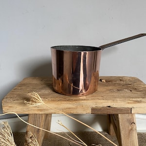 May include: A polished copper saucepan with a straight, dark metal handle extending to the right. The cylindrical pot features a reflective, warm metallic exterior and a dark interior lining, showcasing a clean, simple design for kitchen use.