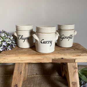 May include: Three cream-coloured ceramic spice jars with lids and handles, labelled "Thym," "Curry," and "Girofle" in black script. The jars sit on a rustic wooden surface, evoking a vintage or farmhouse style.