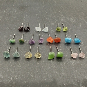 May include: Assortment of 20 pairs of gemstone earrings. Each pair features a unique, irregular shaped gemstone on a silver post. The gemstones are in various colors, including green, blue, red, purple, and brown.