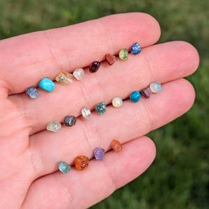Può includere: Una collezione di piccole pietre preziose grezze multicolori di varie forme e consistenze, tra cui sfumature di blu, rosso, arancione, verde, giallo e bianco trasparente, disposte sul palmo di una mano per mostrarne le caratteristiche.