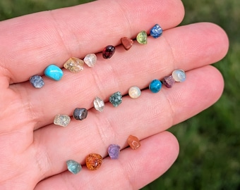May include: A collection of small, multi-colored raw gemstones in various shapes and textures, including shades of blue, red, orange, green, yellow, and clear white, displayed across the palm of a hand to show their individual characteristics.