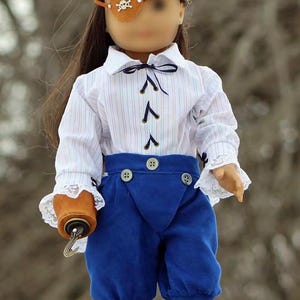 May include: A doll wearing a white and blue striped shirt with a blue bow tie, blue pants with white lace trim, and a brown leather eye patch with a silver skull and crossbones design. The doll is holding a brown leather pouch with a metal hook.