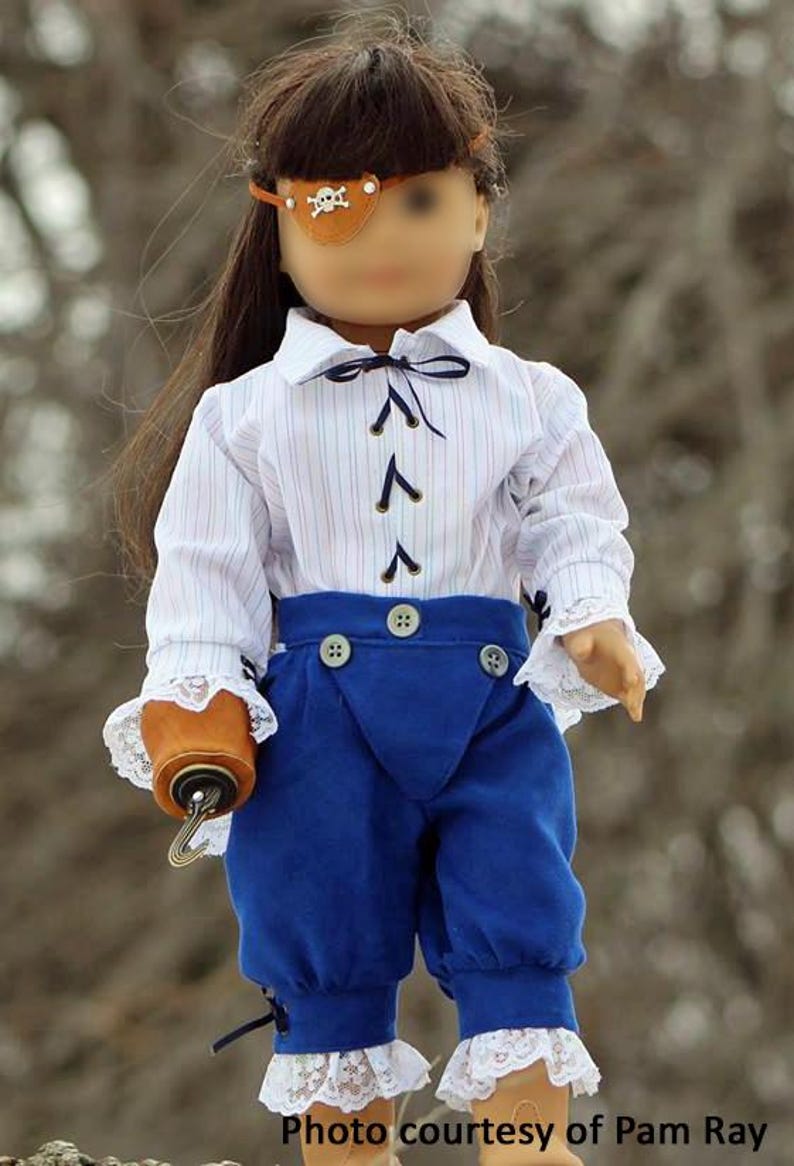 May include: A doll wearing a white and blue striped shirt with a blue bow tie, blue pants with white lace trim, and a brown leather eye patch with a silver skull and crossbones design. The doll is holding a brown leather pouch with a metal hook.