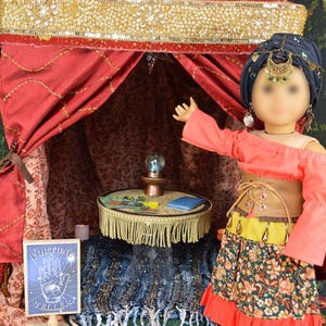 May include: A doll wearing a red and green outfit stands in a red tent with gold trim. The tent has a small table with a crystal ball and a sign that says "Fortune Teller".