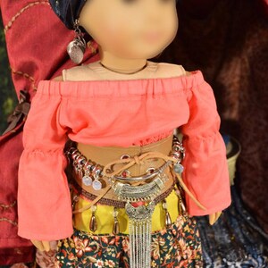 May include: A doll wearing a pink off-the-shoulder top, a brown leather belt with silver accents, a yellow and green skirt with floral print, and a silver chain necklace with a pendant. The doll is wearing a blue turban with silver embellishments and silver earrings.