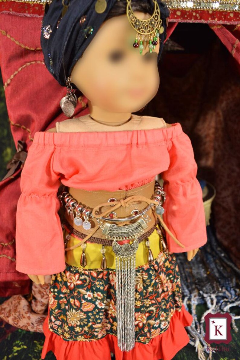 May include: A doll wearing a pink off-the-shoulder top, a brown leather belt with silver accents, a yellow and green skirt with floral print, and a silver chain necklace with a pendant. The doll is wearing a blue turban with silver embellishments and silver earrings.