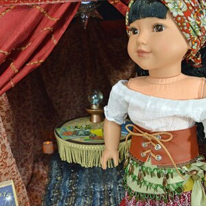 May include: A doll wearing a white off-the-shoulder blouse, a brown leather belt with laces, a green and gold beaded skirt, and a red and white headscarf. The doll is standing in a red and gold tent with a small table and a crystal ball.