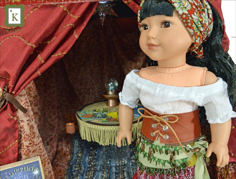 May include: A doll wearing a white off-the-shoulder blouse, a brown leather belt with laces, a green and gold beaded skirt, and a red and white headscarf. The doll is standing in a red and gold tent with a small table and a crystal ball.