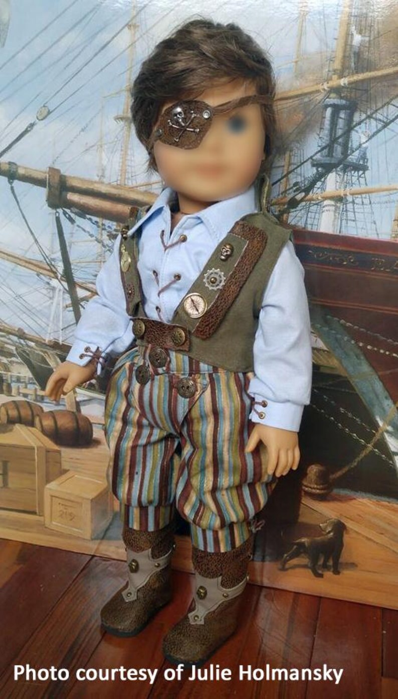 May include: A doll wearing a blue shirt, brown vest, striped pants, and brown boots. The doll has brown hair and is wearing an eye patch with a skull and crossbones design.