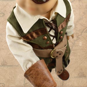 May include: A child's pirate costume with a green vest, brown belt, and a brown leather hook hand. The vest has brown ribbon accents and copper buttons. The child is wearing a brown hat and an eye patch with a skull and crossbones design.