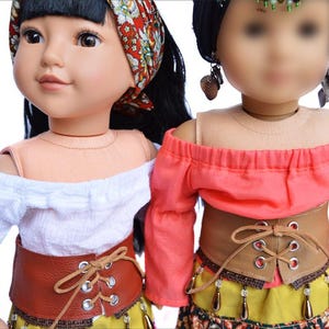 May include: Two dolls wearing off-the-shoulder tops and brown leather belts with laces. The doll on the left is wearing a white top and the doll on the right is wearing a coral top. Both dolls are wearing yellow skirts with beaded accents.