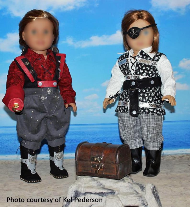 May include: Two dolls dressed as pirates, one with a red shirt and black vest, the other with a white shirt and black vest with skull and crossbones pattern. Both dolls are wearing grey pants and black boots. A wooden treasure chest is in the foreground.
