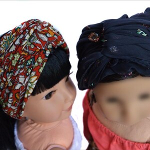 May include: Two dolls wearing headbands. One doll has black hair and is wearing a red and white floral headband. The other doll has brown hair and is wearing a dark blue headband with glitter.