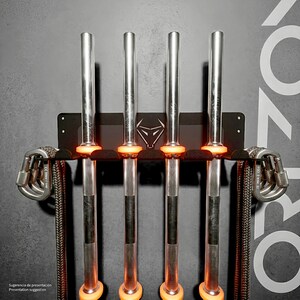 May include: A black metal wall-mounted rack featuring four vertical slots for barbells, each with bright orange protective rings. The central plate displays a stylised bull head logo. Two silver carabiners with thick grey ropes hang from the sides.