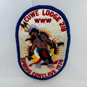 May include: Embroidered patch with a blue border, featuring text and an illustration. The text reads "CUWE LODGE 218" and "SPRING CONCLAVE 1974". The illustration depicts a figure with a feathered headdress and a sunburst design.
