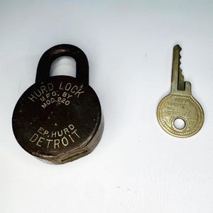 May include: A vintage Hurd Lock with its key. The lock is round, dark brown, and has the text "HURD LOCK MFG. BY MOD. 220 E.P. HURD DETROIT" printed on it. The key is a light gold color and has the text "SECURITY FOR AMERICA" printed on it.