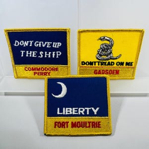 May include: Three embroidered patches with patriotic themes. One patch features the text "DON'T GIVE UP THE SHIP" and "COMMODORE PERRY" on a navy blue background with a gold border. Another patch displays a coiled snake with the text "DON'T TREAD ON ME" and "GADSDEN" on a yellow background. The third patch has a crescent moon and the word "LIBERTY" with "FORT MOULTRIE" on a navy blue background.