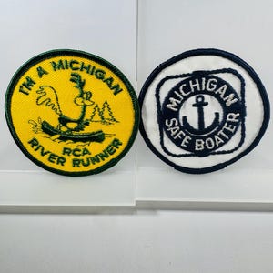 May include: Two round embroidered patches. One is yellow with green text and a cartoon character in a boat. The text reads "I'M A MICHIGAN RIVER RUNNER RCA". The other is white with navy blue text and an anchor. The text reads "MICHIGAN SAFE BOATER".