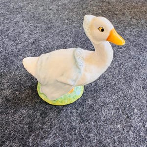 May include: A ceramic goose figurine with a white body, yellow beak, and black eyes. The goose wears a light blue bonnet and stands on a green and yellow base. The figurine is a decorative item.