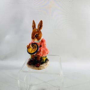 May include: A ceramic figurine of a brown rabbit wearing a pink dress and holding a blue and gold plate. The rabbit has long ears and is standing on a small, round base. The figurine is a decorative item.