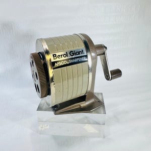 May include: A vintage Berol Giant APSCO pencil sharpener featuring a beige cylindrical body with vertical grooves, a brown rotating handle, and a dark brown dial with multiple holes for different pencil sizes, mounted on a sturdy metal base.