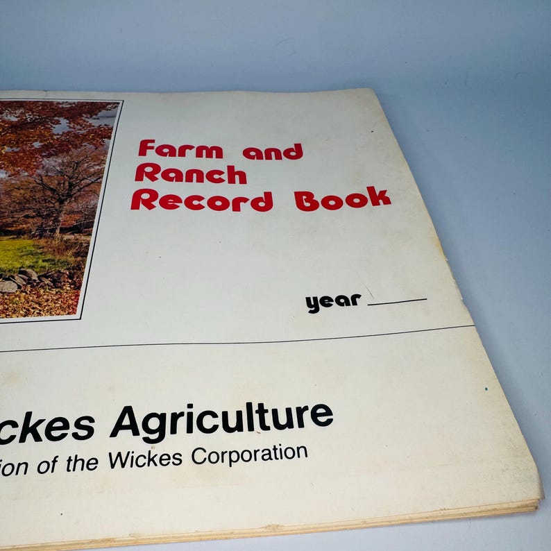 Pu&ograve; includere: Un vecchio "Farm and Ranch Record Book" con testo rosso su una copertina color crema. Il libro include il testo "Wickes Agriculture" e uno spazio per scrivere l'anno. Una foto di paesaggio &egrave; a sinistra.