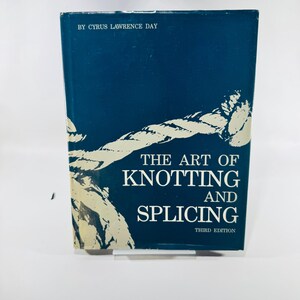 May include: A hardcover book titled The Art of Knotting and Splicing, Third Edition, by Cyrus Lawrence Day. The dark blue cover features a large, cream-colored graphic illustration of a rope tied in a knot across the front surface.