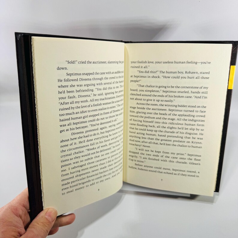 May include: An open book with black covers and cream-coloured pages. The text is in a serif font. The book is held open, revealing text on both sides. The edges of the pages are visible, and the spine is on the left.