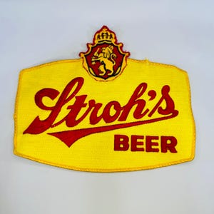 May include: A yellow embroidered patch featuring the red text Stroh's BEER in a script and block font. At the top, a red and yellow crest displays a lion holding a mallet beneath a crown. The patch has a distinct shield-like shape with red borders.