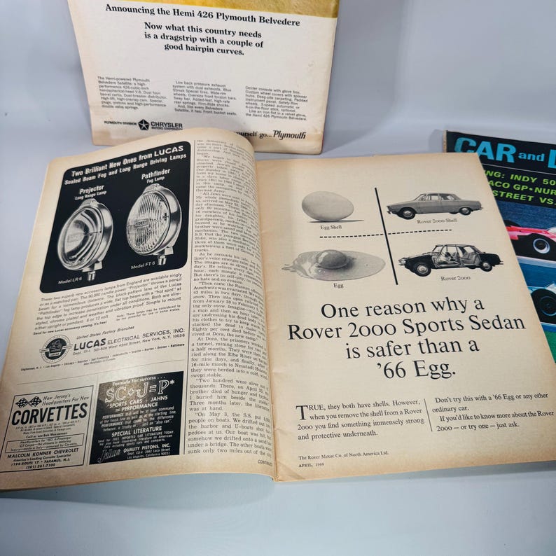 May include: A vintage magazine open at several pages with car advertisements. One advertises the Hemi 426 Plymouth Belvedere. Another features Lucas driving lamps. A third shows a Rover 2000 Sports Sedan with the text "One reason why a Rover 2000 Sports Sedan is safer than a '66 Egg."