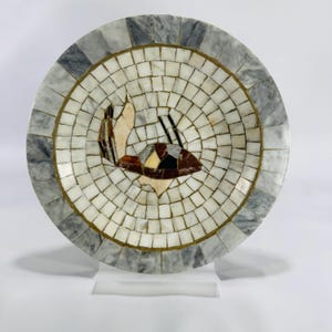 May include: A round decorative plate with a mosaic design. The plate features a central image of a sailboat, crafted from various colored stones. The border is a mosaic of gray and white marble, with a gold-colored trim.