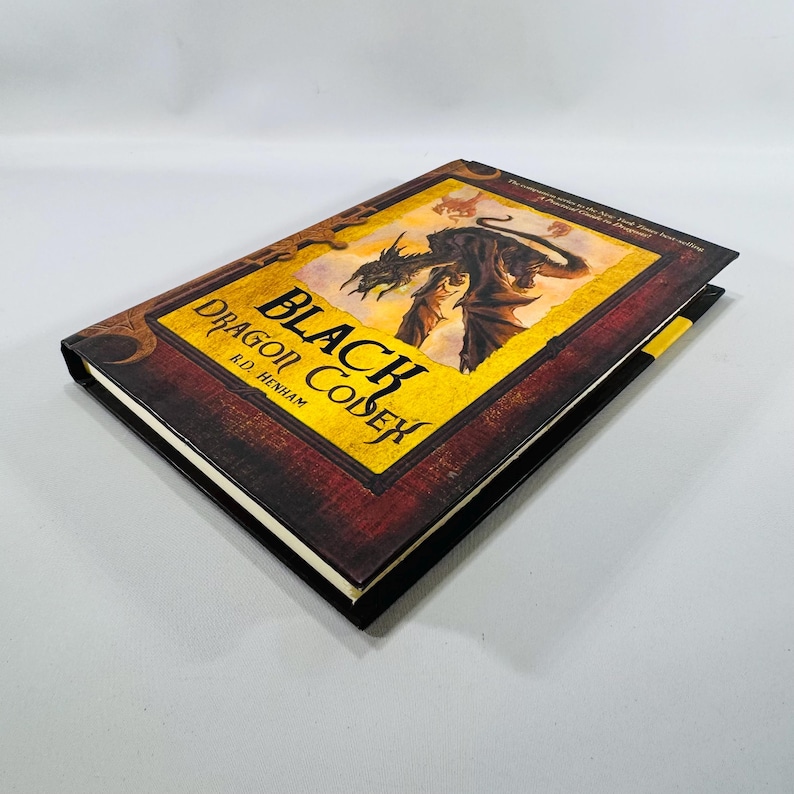 May include: A hardback book titled "Black Dragon Codex" by R.D. Henham. The cover displays a dragon illustration within a yellow and black frame with a red border. The book has a black spine and a yellow bookmark.