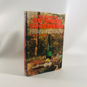 May include: The Complete Outdoor Cookbook book cover with a red title and a photo of a bottle of wine, a knife, eggs, and meat on a wooden cutting board.