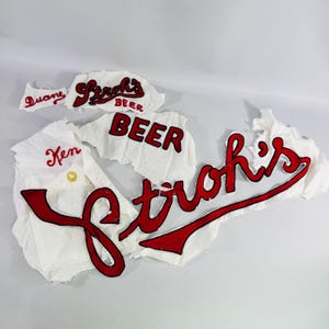 May include: A collection of white fabric patches featuring red embroidered lettering. The patches include the names Duane and Ken, along with the text Stroh's, Beer, and Stroh's Beer. The embroidery is in a bold red script and block font style on white fabric scraps.