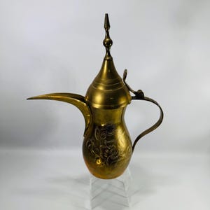 May include: A vintage brass coffee pot with a long spout and a decorative floral pattern. The pot has a handle and a pointed lid, and it is displayed on a clear stand. The brass has a warm, golden color.