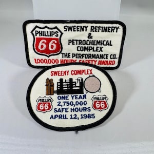 May include: Two embroidered patches with black borders. The rectangular patch reads Phillips 66 Sweeny Refinery & Petrochemical Complex The Performance Co. 1,000,000 Hours Safety Award. The oval patch shows a refinery graphic and Sweeny Complex One Year 2,750,000 Safe Hours April 12, 1985.