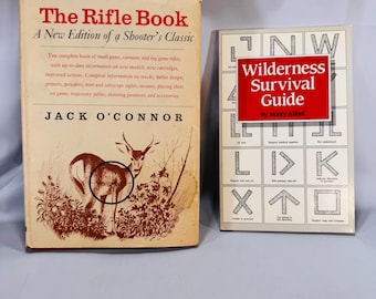 May include: Two vintage books. "The Rifle Book" by Jack O'Connor, featuring a deer illustration, and "Wilderness Survival Guide" by Monty Alford, with a red title box. Both books are on a light blue surface.