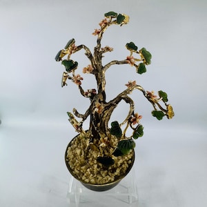 May include: A decorative bonsai tree sculpture. The tree features a gold-colored metal trunk and branches, adorned with green leaves and small pink flowers. The tree sits in a dark-colored pot filled with small, light-colored stones. The sculpture is displayed on a clear stand.
