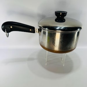 May include: Stainless steel saucepan with a black handle and lid. The pot has a copper-colored bottom and a black knob on the lid. A metal ring is attached to the end of the handle.
