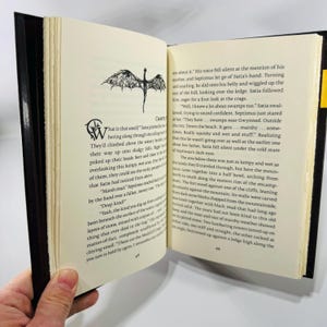 May include: An open book with black covers and yellow page edges, held by a hand. The pages display printed text and a small illustration of a winged creature. The book is likely a novel or a work of fiction.