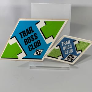May include: Two diamond-shaped patches with the text "TRAIL BOSS CLUB" in black letters. The larger patch has a blue center and green accents. The smaller patch is embroidered with a blue center and green accents. Both have the "DX" logo.