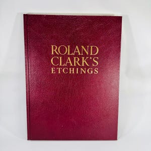 May include: A burgundy-colored book titled "ROLAND CLARK'S ETCHINGS" in gold lettering. The book has a textured cover and a rectangular shape. The book is likely a collection of etchings.