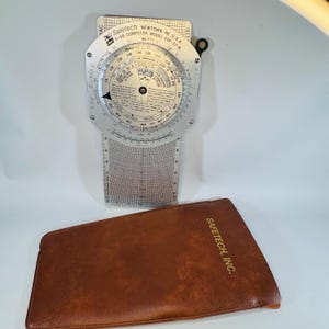 May include: A Safetech E-6B computer model FDF-57-B with a brown leather case. The computer has a circular dial with various scales and measurements. The case has the text "Safetech, Inc." printed on it.