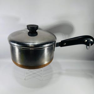 May include: A stainless steel saucepan with a lid and a black handle. The pot has a copper-colored bottom and a black knob on the lid. The handle has a loop at the end. This kitchenware is designed for cooking.