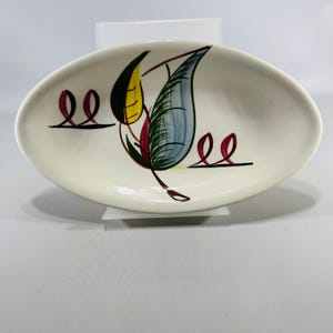 May include: An oval-shaped white ceramic dish featuring a hand-painted design of a central leaf motif in shades of yellow, blue, and green, flanked by two sets of stylized maroon ribbon-like loops on horizontal black lines on either side.