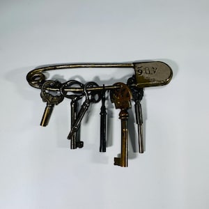 May include: A collection of vintage keys hangs from a large, antique-style safety pin. The pin is a tarnished brass color, and the keys vary in size and design, with some showing ornate details. The number "406" is stamped on the pin's head.