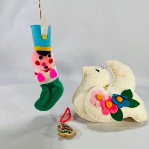 May include: A collection of vintage Christmas ornaments. Includes a felt soldier in a stocking, a white dove with floral accents, and a gingerbread tree. Each ornament has a gold string for hanging.